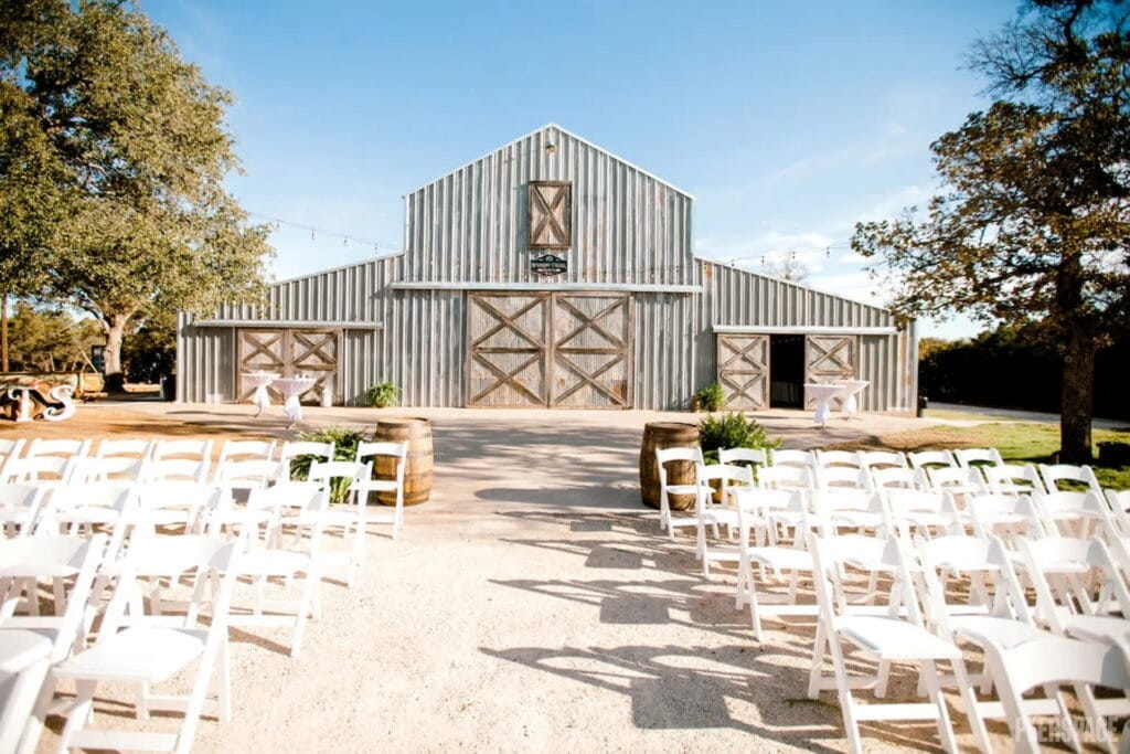 Simple white chairs are arranged for a wedding ceremony outdoors in front of a rustic barn