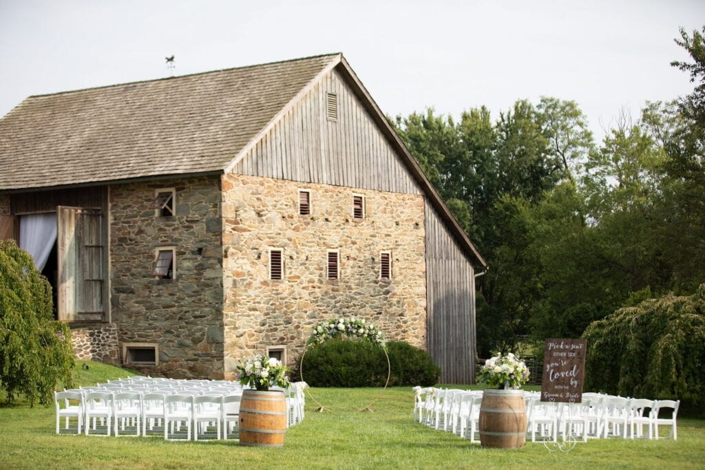 White wedding ceremony chairs line an outdoor aisle by a stone and wood barn
