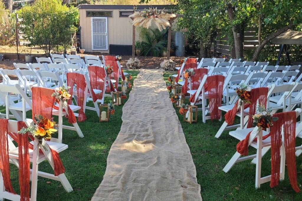 A burlap runner serves as an aisle at an outdoor wedding ceremony decorated with white wooden chairs accented with deep red fabric, and orange and yellow flowers