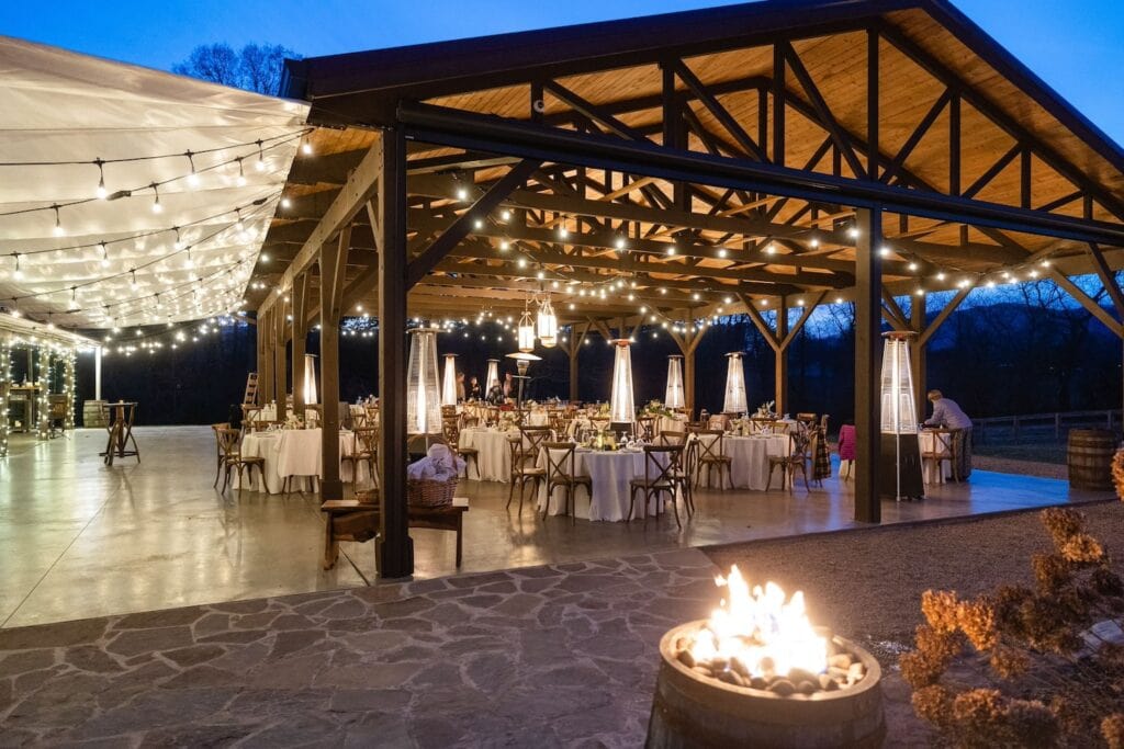 A large outdoor wedding venue is lit up by string lights, outdoor heaters and candles