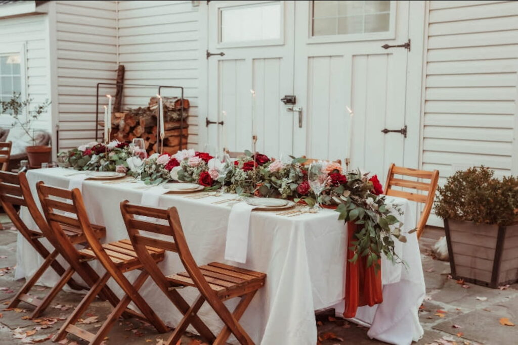 Small reception table set up outside by a farmhouse door and decorated with red, pink and white flowers set in greenery