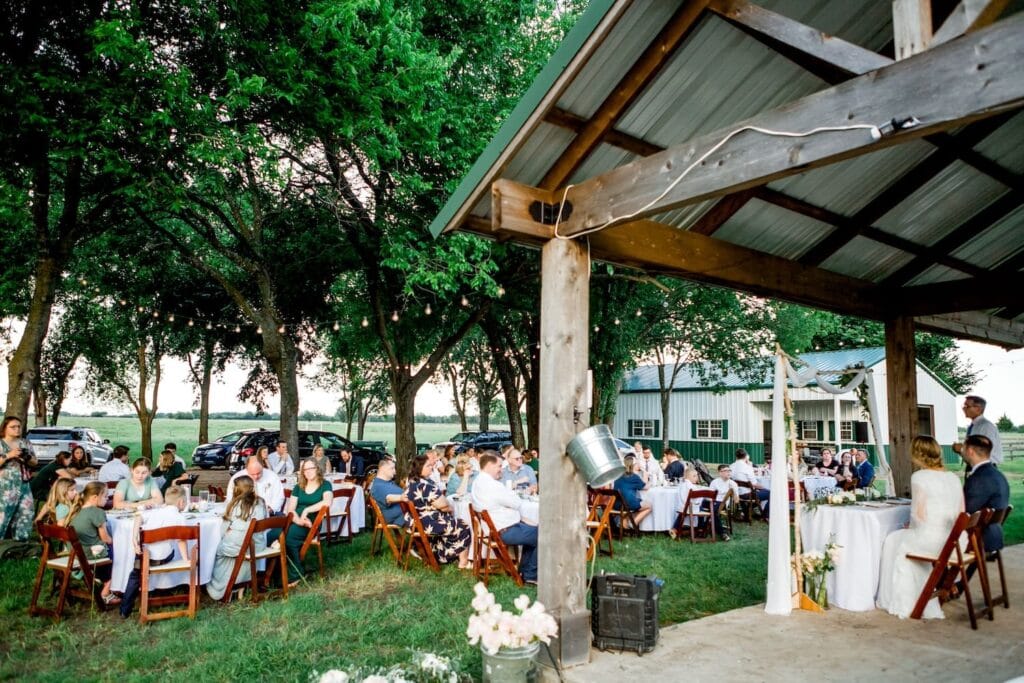An outdoor reception under tall trees strung with string lights next to a barn