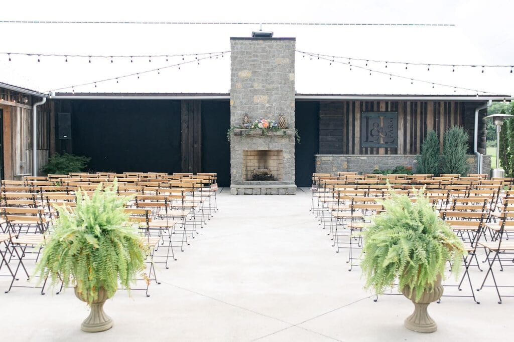 A wedding ceremony is set up by a large outdoor fireplace underneath string lights