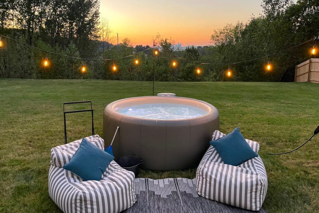 A small hot tub sits in a backyard surrounded by string lights and beanbag chairs with the sun setting in the distance