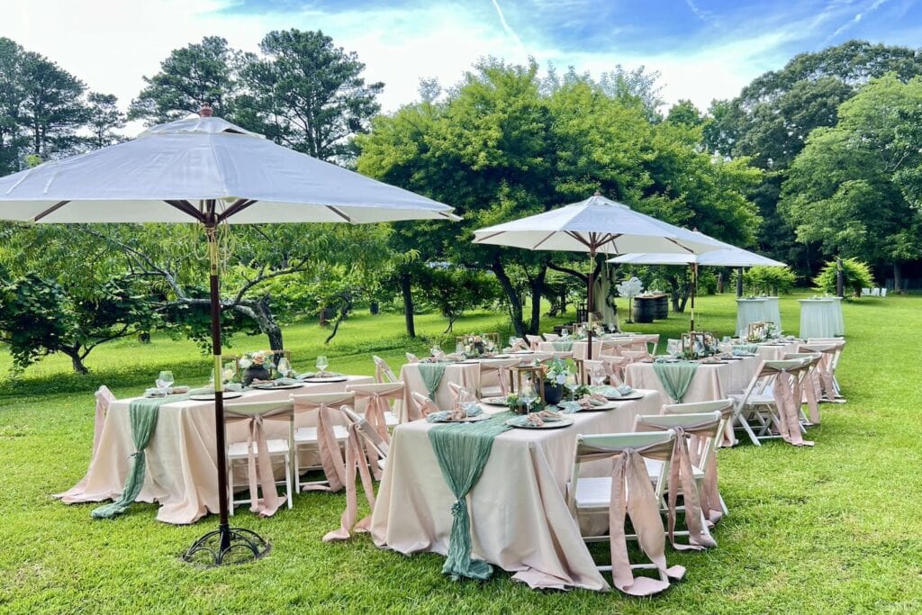 Pink and green linens decorate a casual outdoor venue space set up in the grass under large umbrellas
