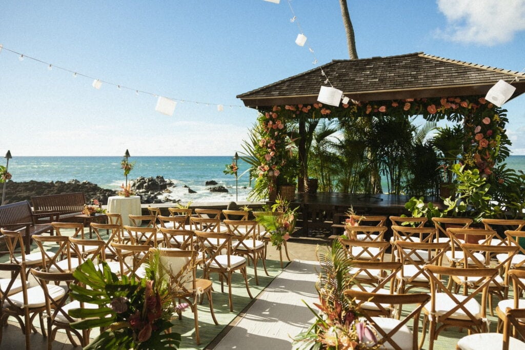 An oceanside gazebo is decorated with pink flowers and palms with wooden chairs set up for a wedding reception