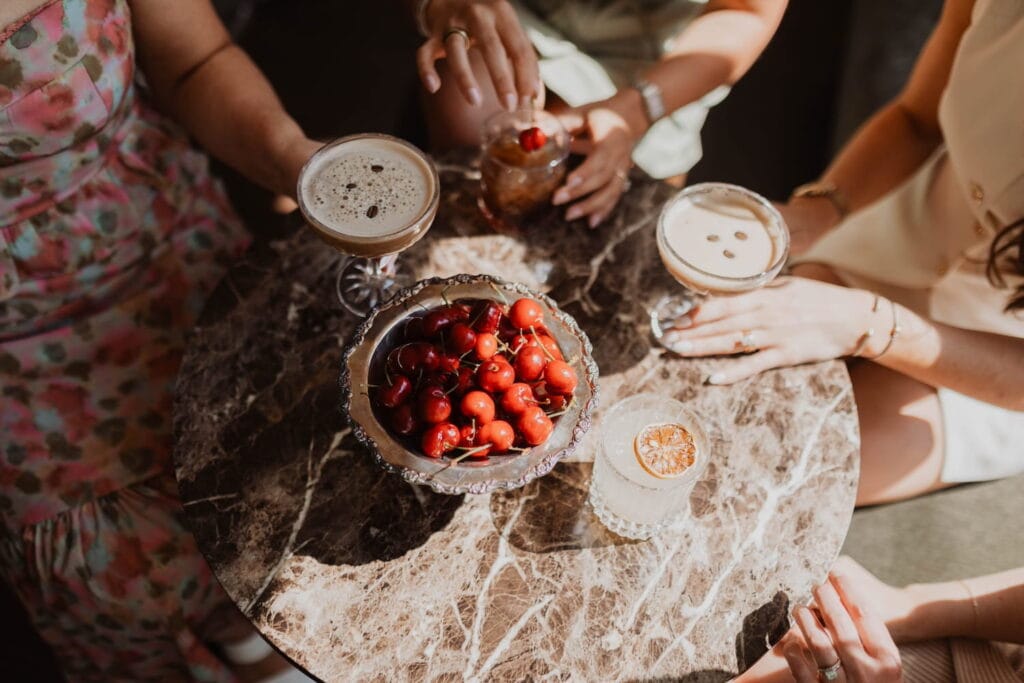 An overhead shot of a marble table in the sunlight with women holding a mixture of cocktails and a bowl of cherries in the middle