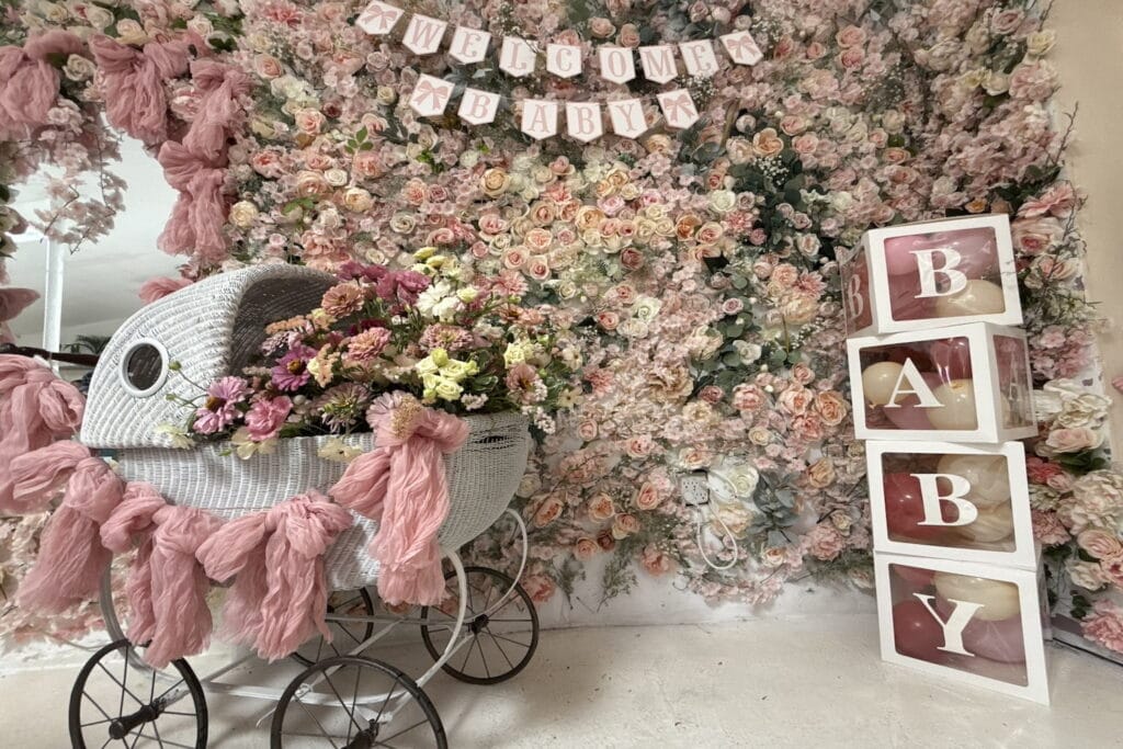 A wicker pram is full of pink flowers and decorated in pink bunting in front of a pink flower wall
