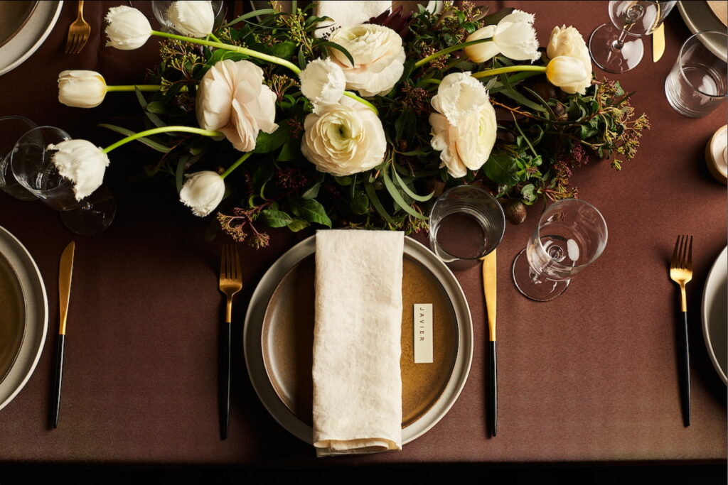 A beautifully simple place setting sits on a table covered in a maroon tablecloth next to gold and black cutlery with a centerpiece of white and green florals