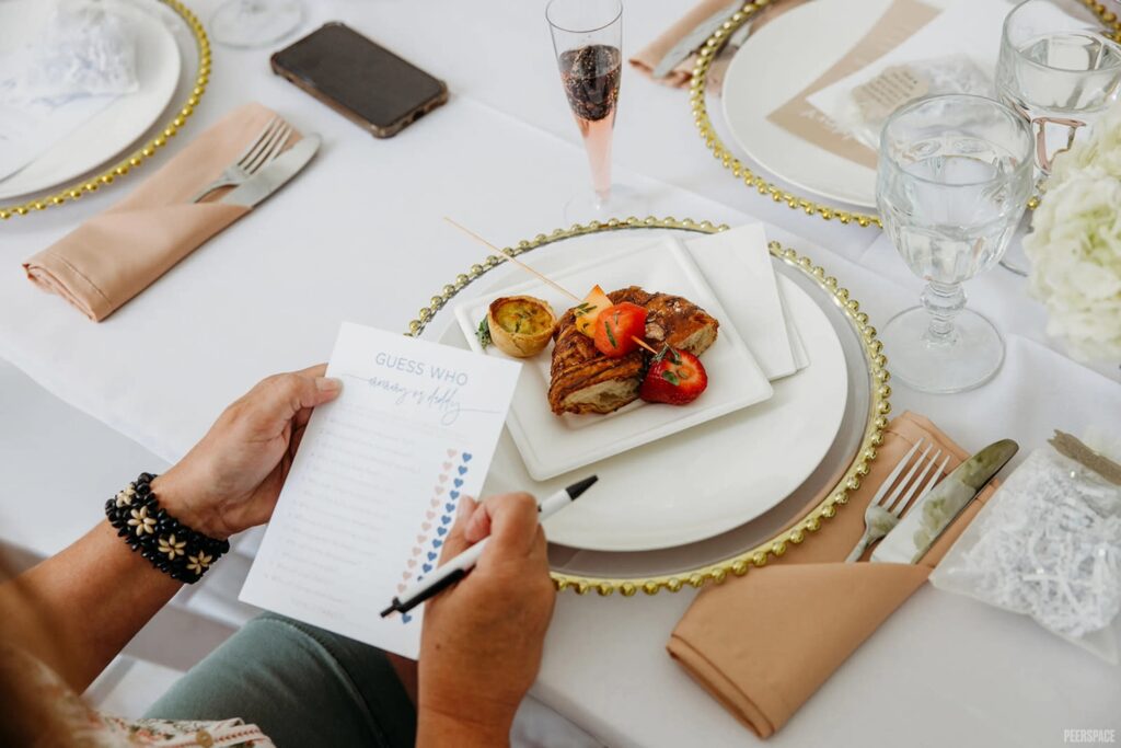 A guest plays a baby shower game while a plate of breakfast foots sits at her place setting