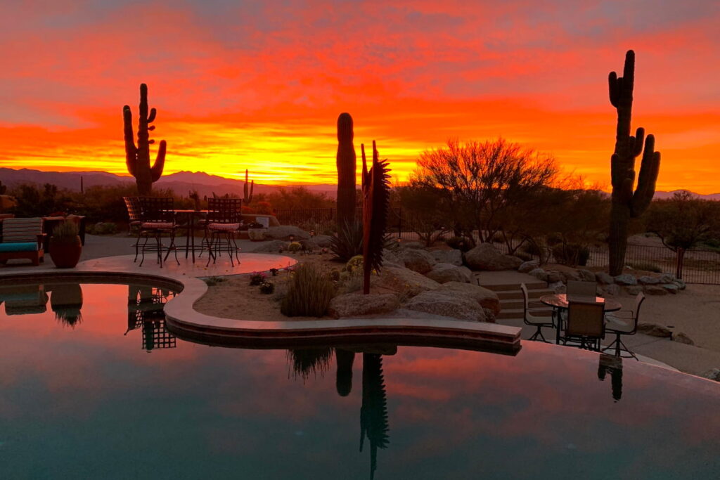 Beautiful pool in backyard of a desert home surrounded by trees and cacti illuminated by a bright red sunset