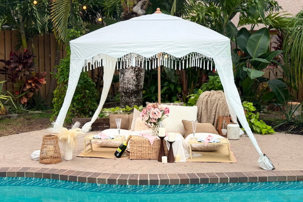 Poolside, boho-themed canopy with a picnic setup underneath styled with pillows, low tables, champagne, flowers and candles