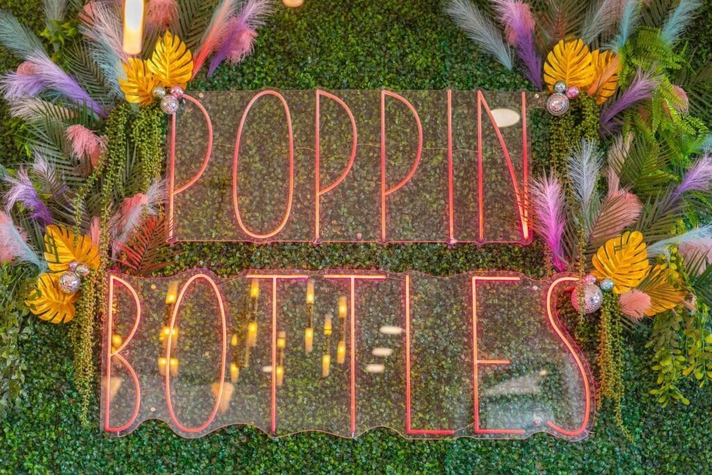 Close up of a neon sign hung on a wall of greenery that says, "Popppin Bottles"