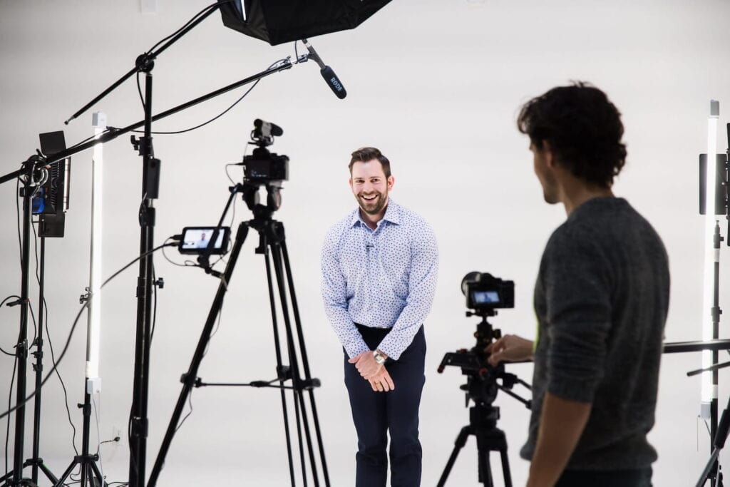 A man stands in a production studio in front of cameras and lighting laughing with a cameraman