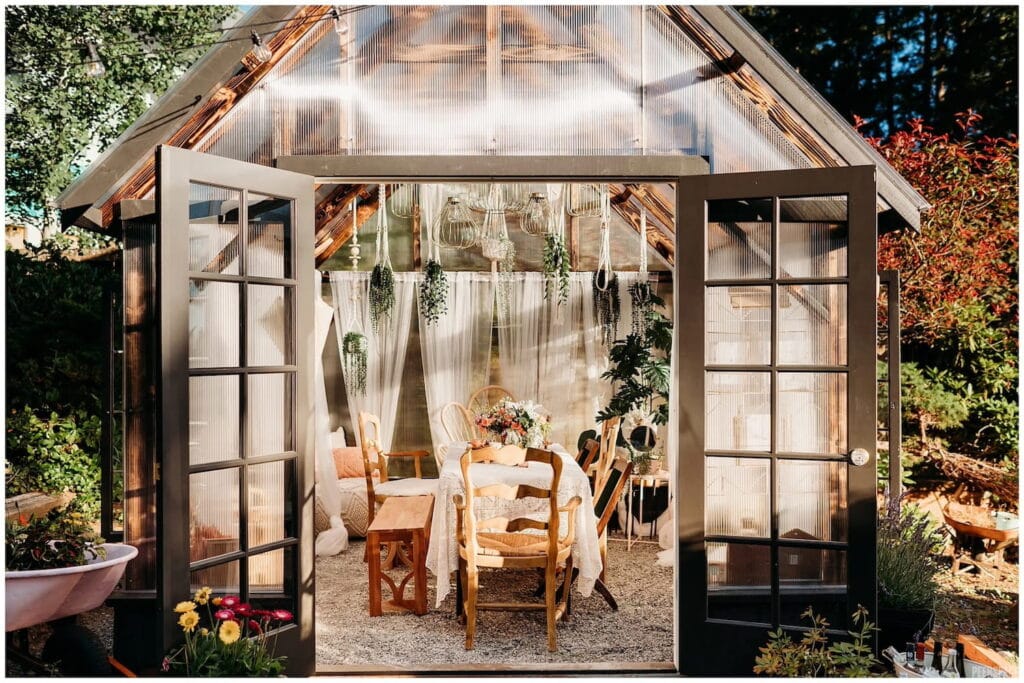 Small dining table set up inside a greenhouse with greenery hanging from ceiling