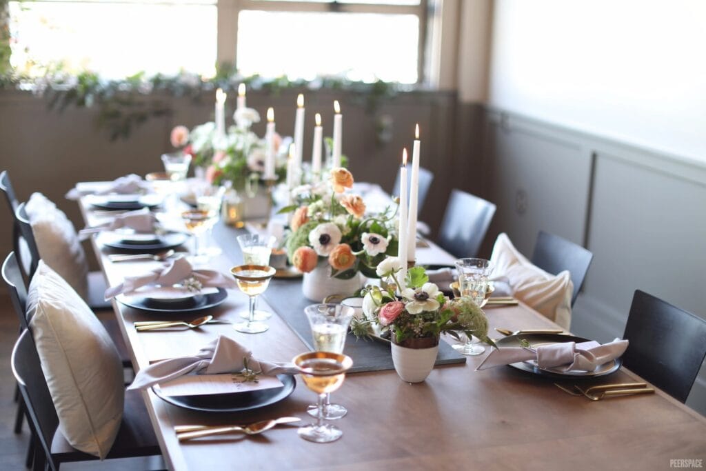 A quaint guest table with low-lying flower arrangements and taper candles is lit up by sunlight from a large window in a neutral-colored space