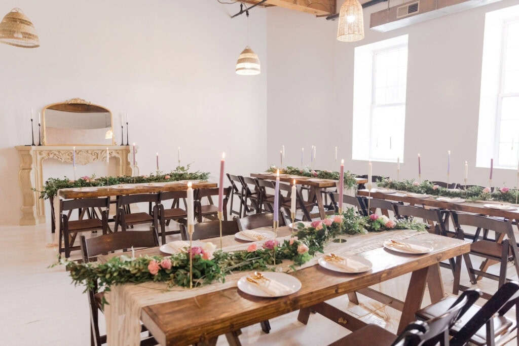 Plain white room with two windows and an ornate off white fireplace mantel set up for a small reception with wooden tables and chairs each decorated with tall taper candles and greenery garland