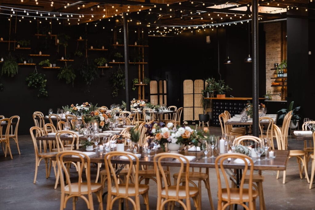 Wood tables and chairs are decorated with low flower arrangements in a dark event space with twinkling lights hanging from the ceiling and a variety of plats hanging on the far wall