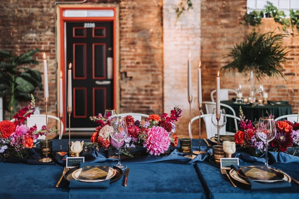 Richly-hued floral arrangements decorate a table clothed in navy in a space with exposed brick walls and a black and red door