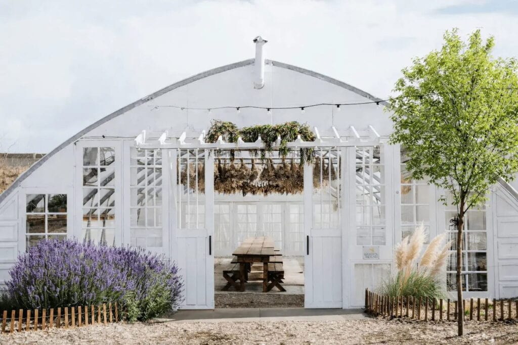 Exterior shot of a white greenhouse with doors open to show long dining table and dried flowers hanging from ceiling
