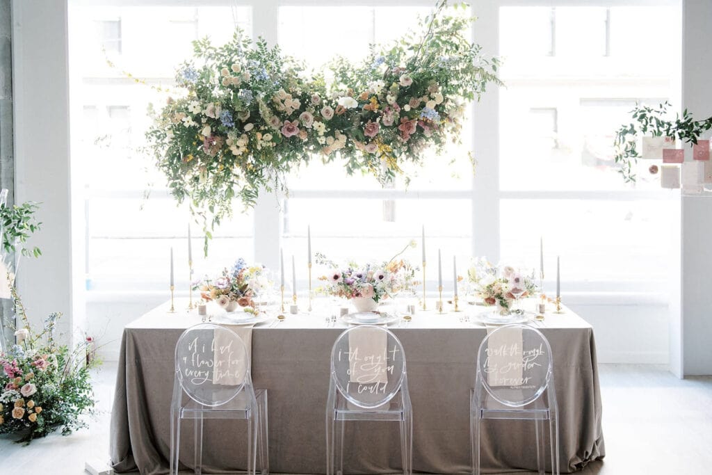 A hanging flower arrangement is suspended above a sweetly-decorated reception table by a large window in bright, white venue