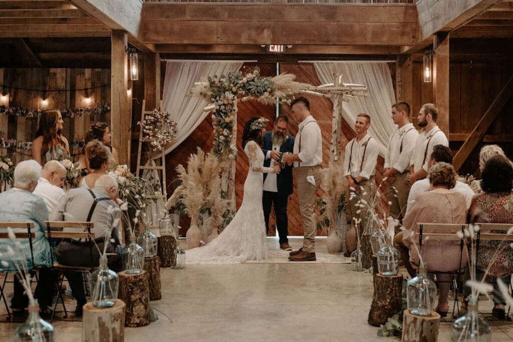 A bride and groom exchange vows at a bohemian-inspired altar constructed of birch tree branches and decorated with flowers and feathers in a rustic barn setting surrounded by friends and family