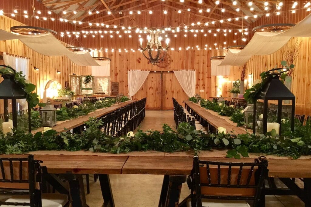 A rustic cabin is set up with long guest tables covered in centerpieces made of greenery and lanterns underneath bistro lights and gauze draperies