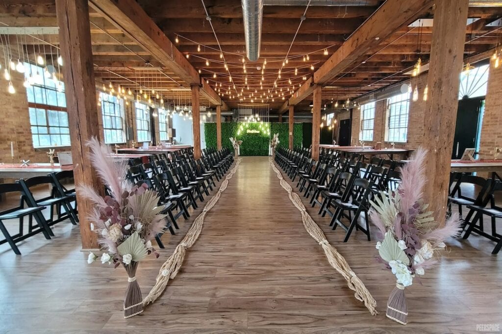 String lights hang above black ceremony chairs that line a rustically decorated wedding aisle in a wood and brick venue