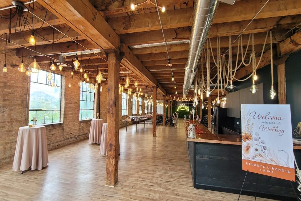 A variety of lightbulbs of varying sizes hang from wooden beams in a rustic event space with exposed brick walls and visible ductwork
