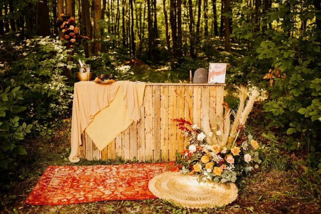 A rustic wooden bar is set up outside in a wooded area decorated with autumnal flowers and layered area rugs