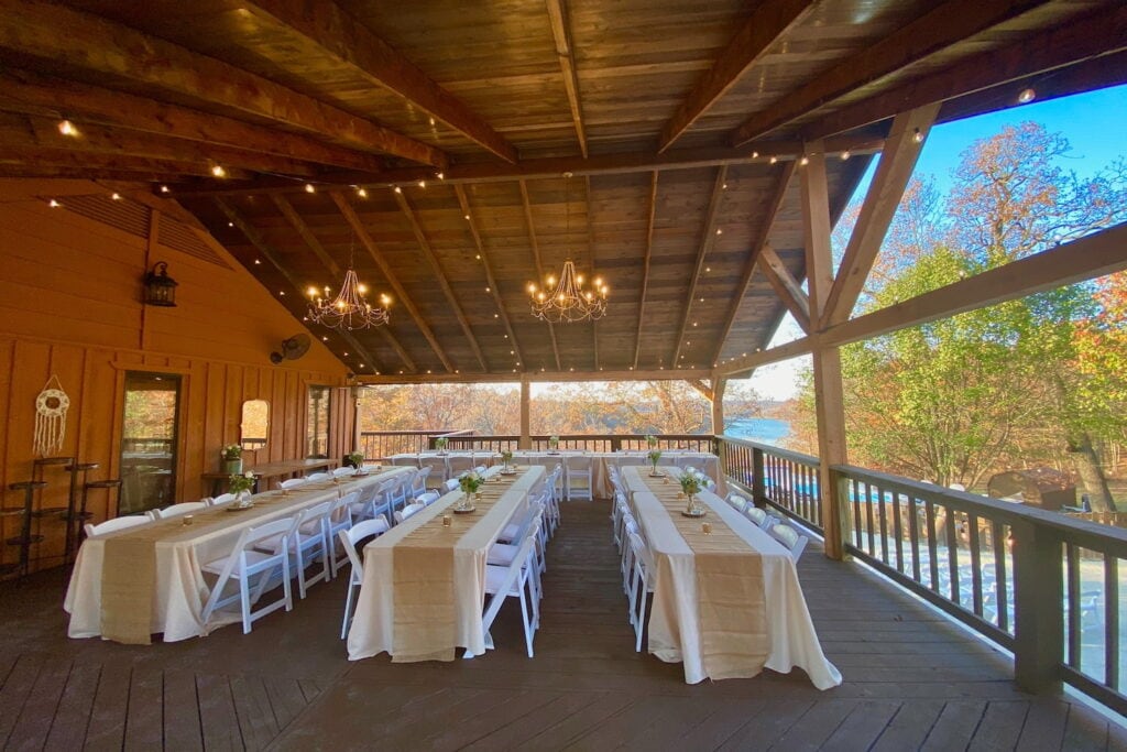 Long reception tables draped in white tablecloths and tan runners sit beneath chandeliers on an expansive, covered outdoor deck