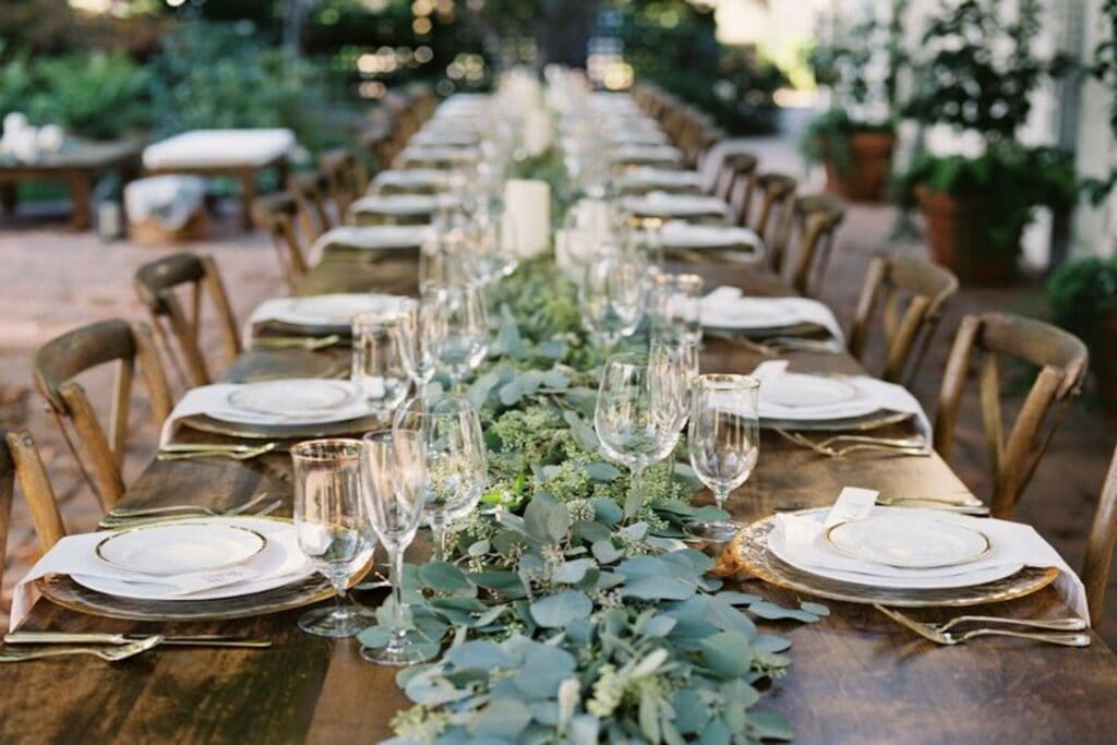 Rustic wooden reception table with matching wooden chairs set with gold and white plates, clear glassware and greenery running down the middle