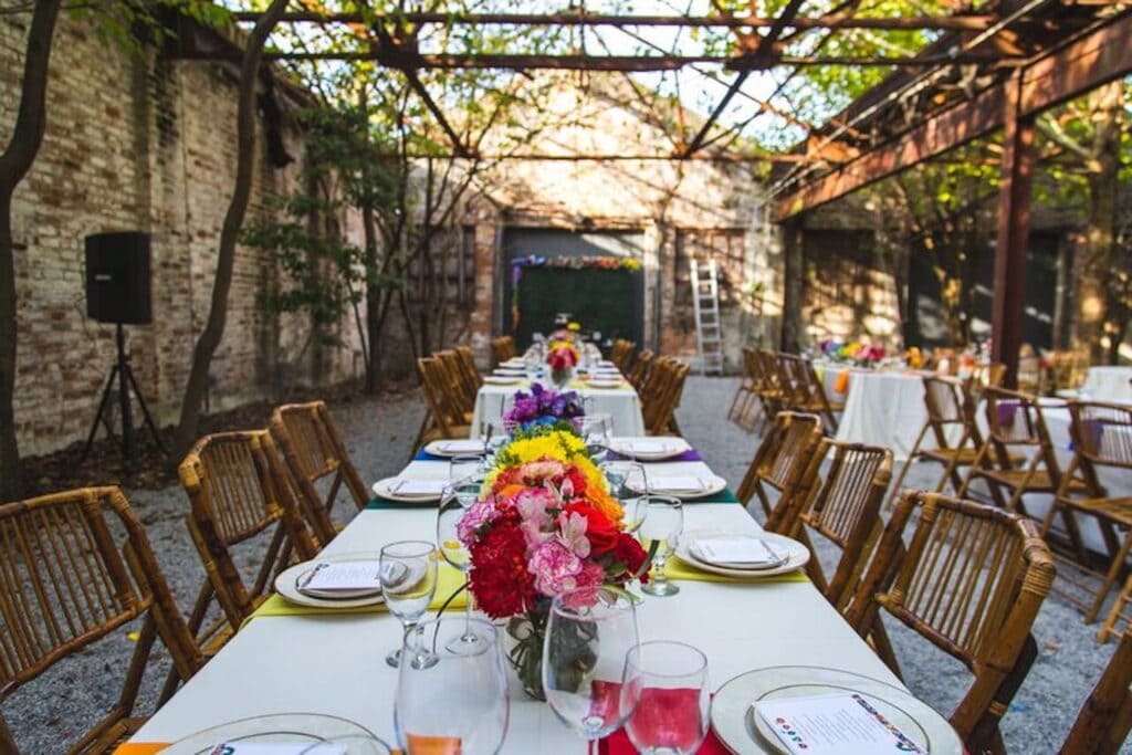 Wedding reception in rustic, open-air building with brick walls and exposed beams set with long tables decorated with brightly-colored flowers