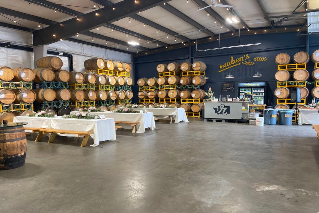 Long picnic tables in a brewery filled with barrels are set up for a reception
