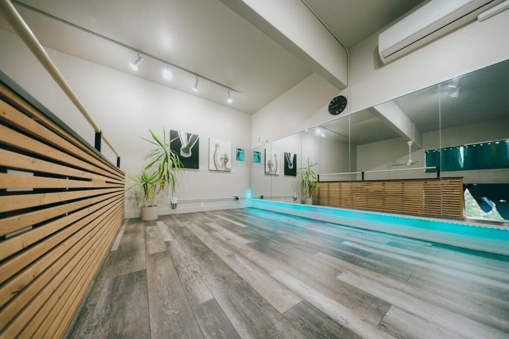A small dance studio with wide-plank wood floors, white walls, and an aqua backlight behind a wall of mirrors