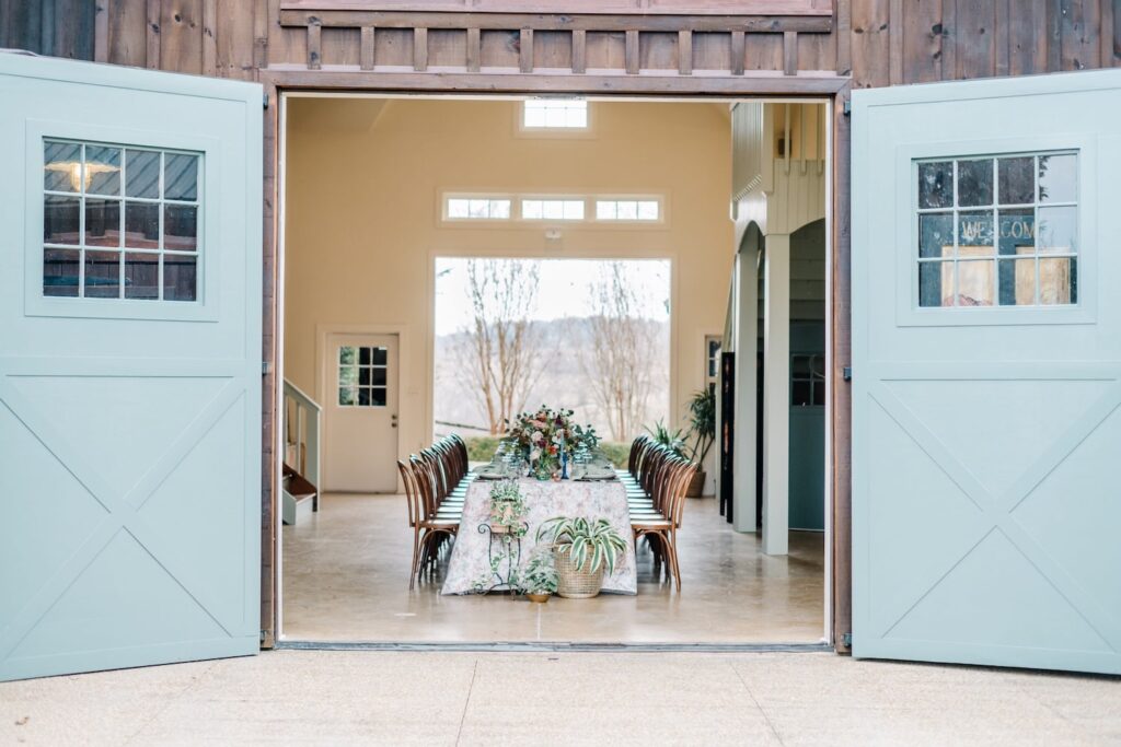 Large baby blue barn doors open to show a long reception table decorated in a floral tablecloth and richly-colored flowers
