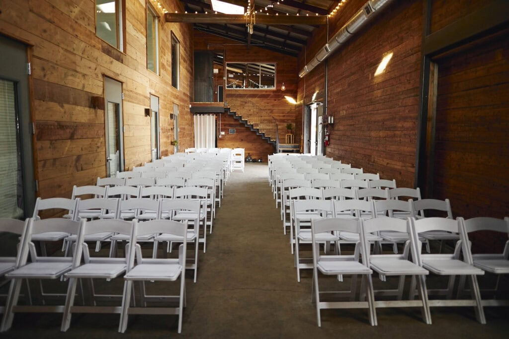 A simple wedding ceremony setup of white chairs in a narrow barn-type structure with wide-plank wood walls and high ceilings