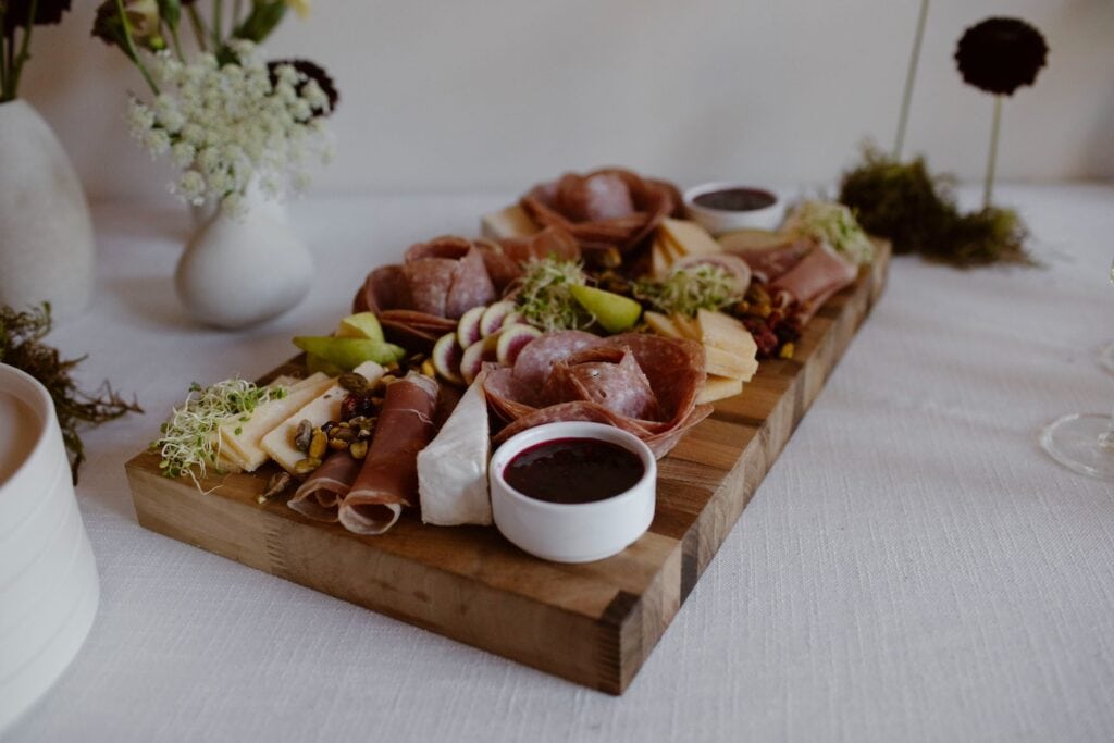 A small selection of cheese and cured meat on a wooden tray surrounded by small vases