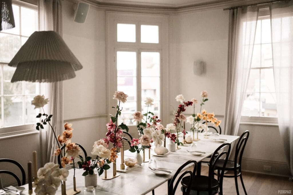 A long table is set with small vases of flowers of all sizes in a bright, white dining room with big windows