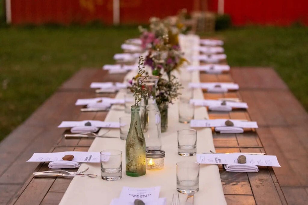 A simple, wooden picnic table is decorated with small flower arrangements in glass bottles in a grassy area