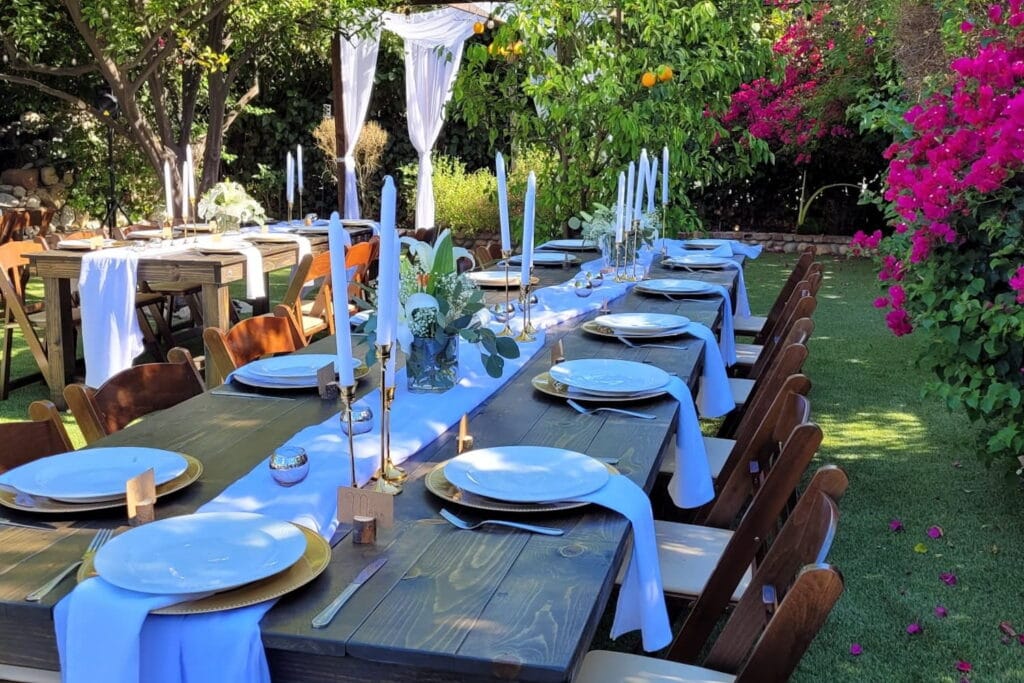 A simply-decorated reception table sits in the shade of a sunny and lush backyard