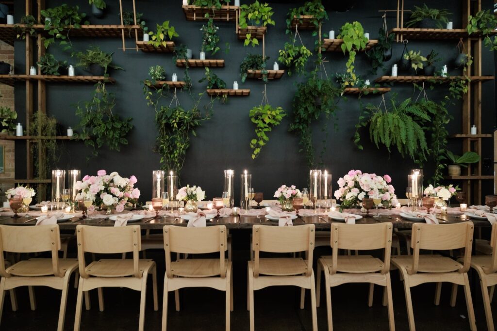 A rich, blue wall full of plants is the backdrop for a simply designed wedding reception with dark tables decorated in pink and white flowers along with taper candles and light-colored chairs.