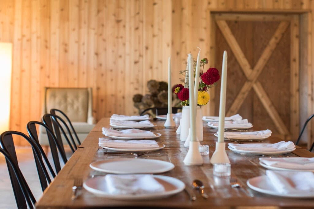 A simple, understated dining table with black metal chairs, white plates and small vases of bright flowers next to tall candles in a wood paneled event space