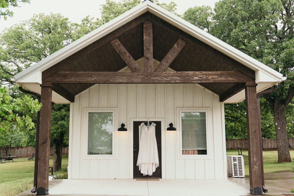 A wedding dress hangs on the outside doorway of a small cottage surrounded by trees