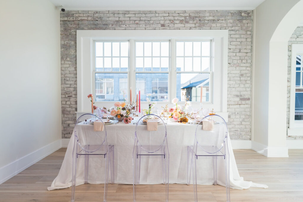 Clear chairs sit around a small reception table decorated with tall candles and colorful flowers in a bright room with whitewashed brick walls and big white windows