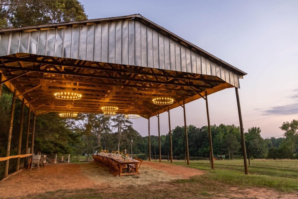 A long reception table is set up for a small gathering underneath a metal pergola lit by large, round chandeliers overlooking a farm