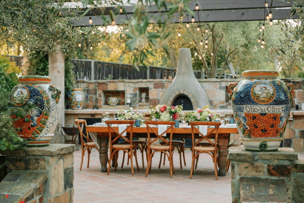 Small reception table set up outside by a chiminea and decorated with white linens, red, white and pink floral arrangements, and blue accents