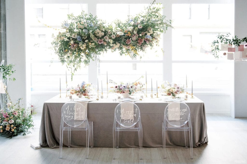 An intimate guest table sits under an elaborate hanging flower arrangement in a brightly-lit reception space