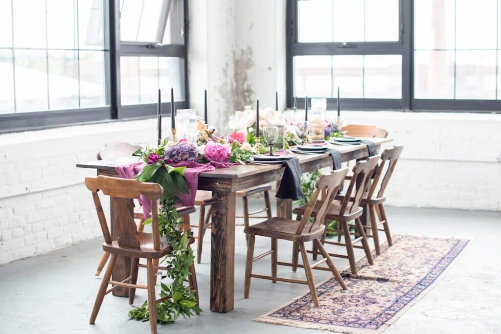 A simple wood table decorated with greenery, pink and purple flowers, and tall taper candles sits in an industrial space with white brick walls and large black windows