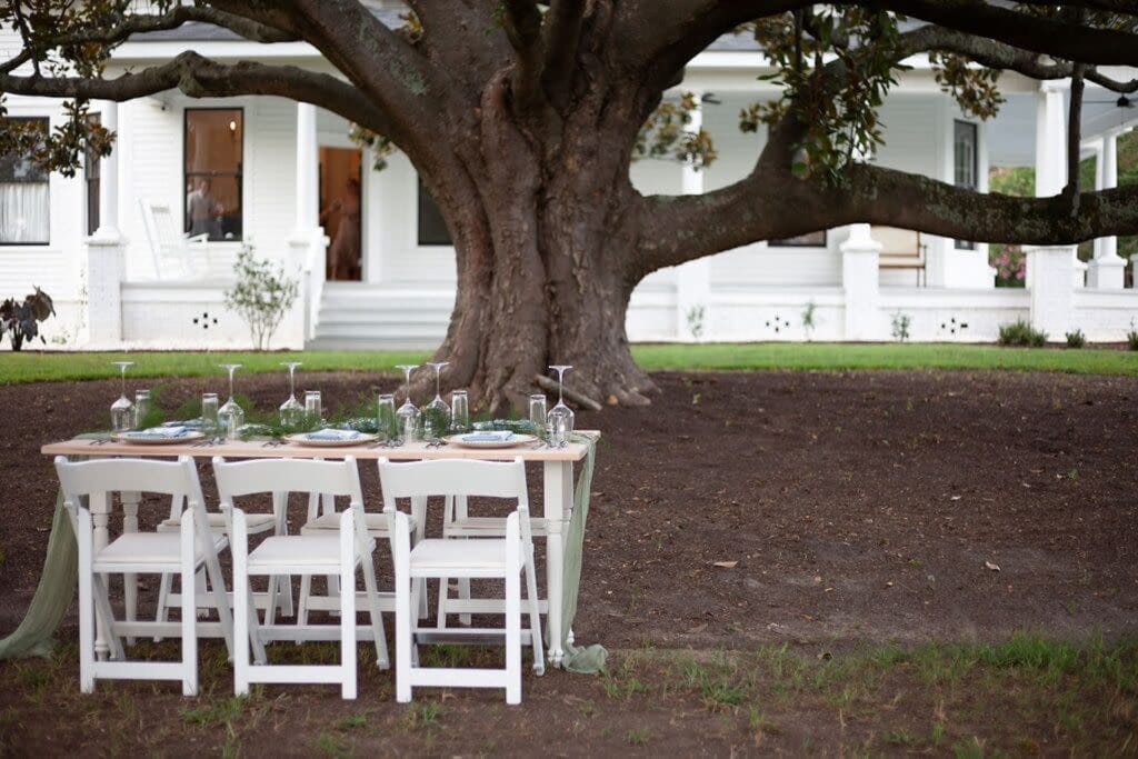 A small dining table set up with greenery under a large tree by a white mansion